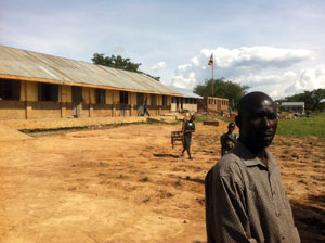Teacher Abraham with the new roofs for local school classrooms from Pilgrim and government built blocks in the background. 